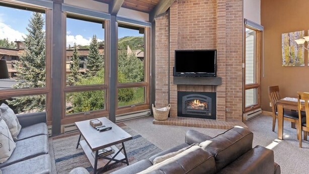 A living room with a fireplace, large windows, and a TV. - The Lodge at Steamboat A301 Upscale condo with balcony, pool, hot tubs, & fireplace - walk to lifts (Steamboat Springs)