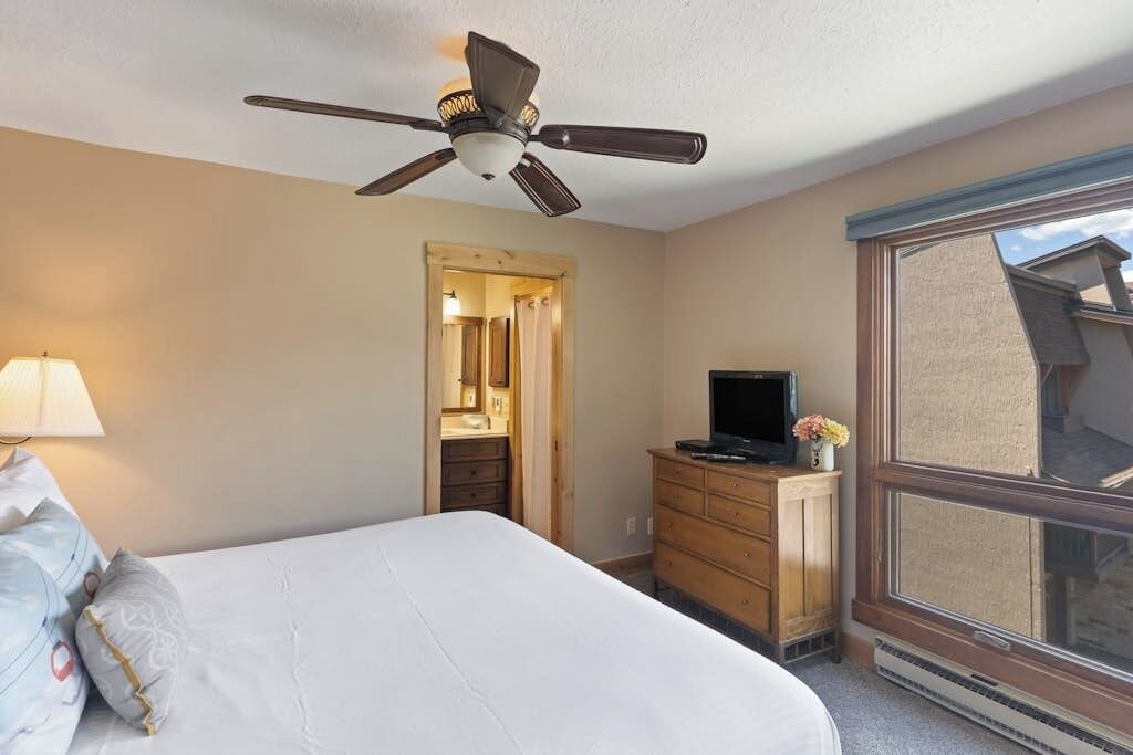 A hotel room with a bed, a ceiling fan, a television, a dresser, and a window.