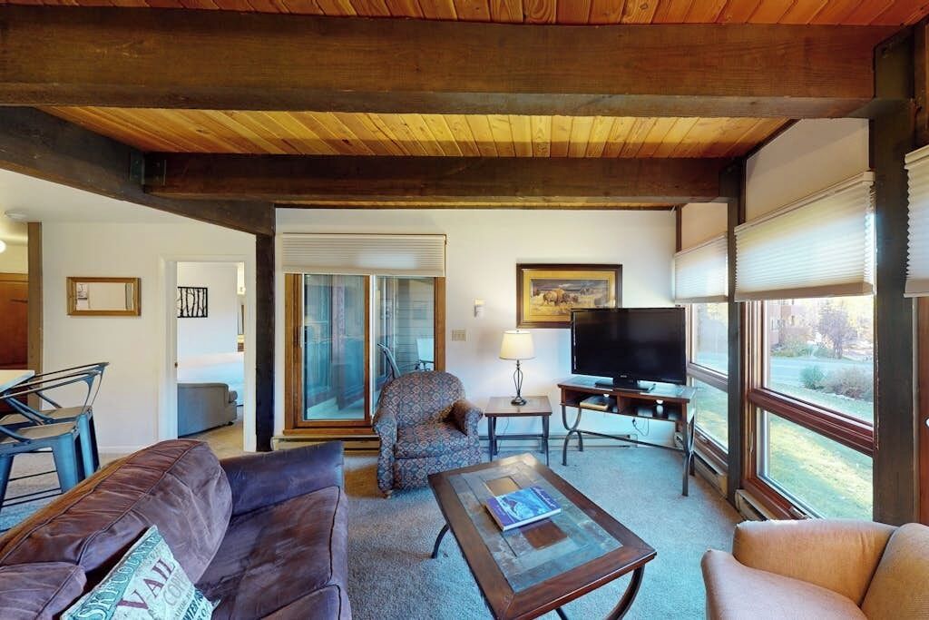 Un salon avec un plafond en bois, un canapé, un fauteuil à motifs, une table basse, un téléviseur et une fenêtre avec des stores.