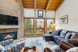 A living room with a fireplace, large windows, a sofa, and a television. - Luxurious mountain condo with pool, hot tubs & patio - near Steamboat ski lifts (Steamboat Springs)