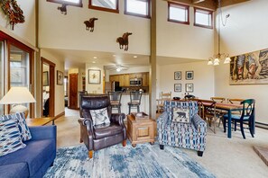 A spacious living room with a high ceiling, a dining area with wooden furniture, and a kitchen in the background. - Luxurious mountain condo with pool, hot tubs & patio - near Steamboat ski lifts (Steamboat Springs)