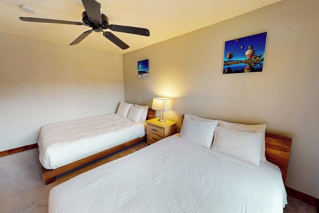 A hotel room with two beds, a ceiling fan, a nightstand with a lamp, and two wall art pieces.