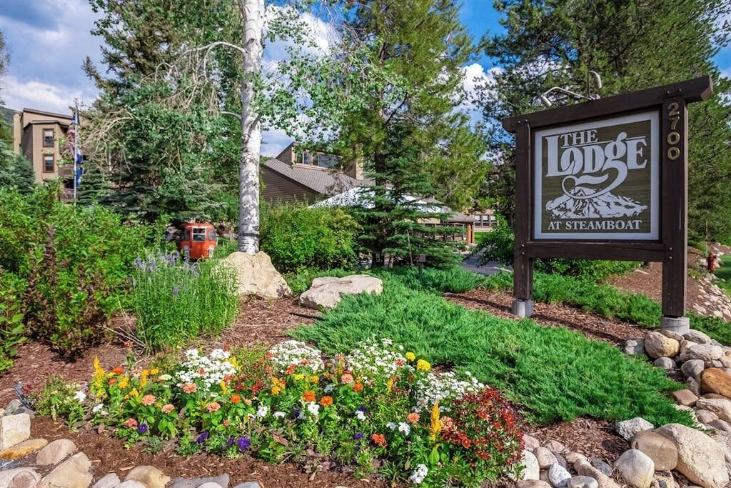 Skilt for 'The Lodge at Steamboat' omgivet af en anlagt have med forskellige planter og et mindre rødt køretøj.