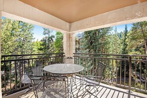 Ein Balkon mit rundem Tisch und Stühlen, umgeben von einem Metallgeländer, mit Blick auf eine bewaldete Gegend.
