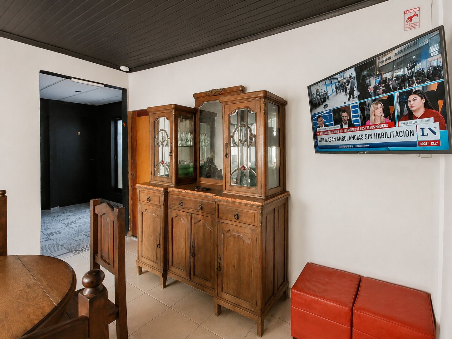 Ein Zimmer mit einem Holzschrank, einem roten Hocker und einem Fernseher, der eine Nachrichtensendung zeigt.