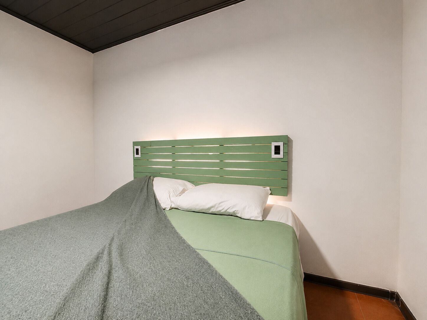 A single bed with a green headboard and white pillows in a room with plain walls and a wooden floor.