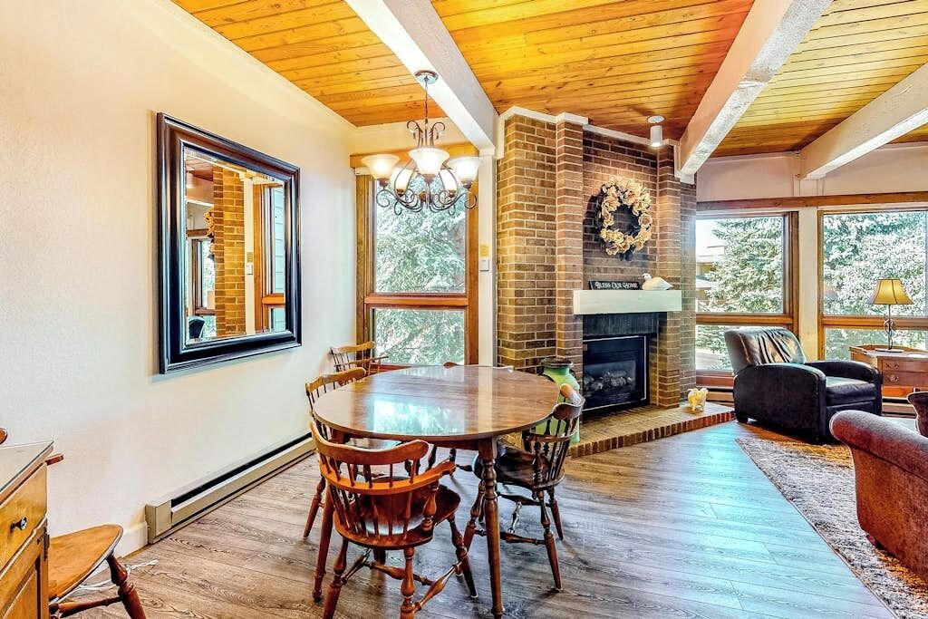 A dining area with a wooden table and chairs, a brick fireplace, and large windows.