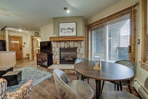 A living room with a stone fireplace, a dining table with chairs, a television, and a kitchen area in the background. - Keystone Condo w/Mountain Views, Shared Pool/Hot Tub - Near Ski Lift/Restaurants (Keystone)