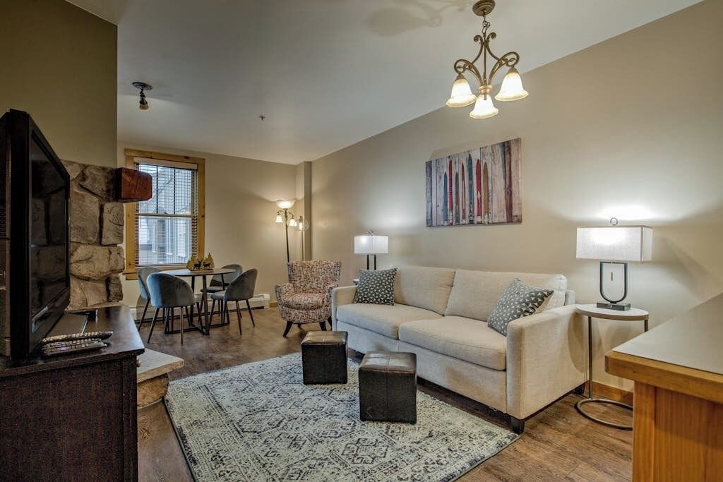 A living room with a sofa, ottomans, a coffee table, a TV, a fireplace, a dining area, and a chandelier.