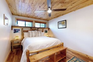 Ein Schlafzimmer mit Holzbalkendecke, einem Bett mit weißer Bettwäsche, einer Holzbank, einem Nachttisch und einem Deckenventilator.