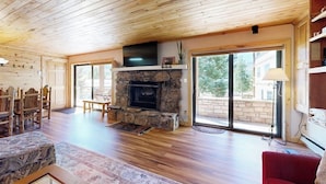 A spacious living room with a stone fireplace, wooden ceiling, and large sliding glass doors leading to an outdoor area. - Charming condo with mountain views, washer/dryer, & shared hot tub (Crested Butte)