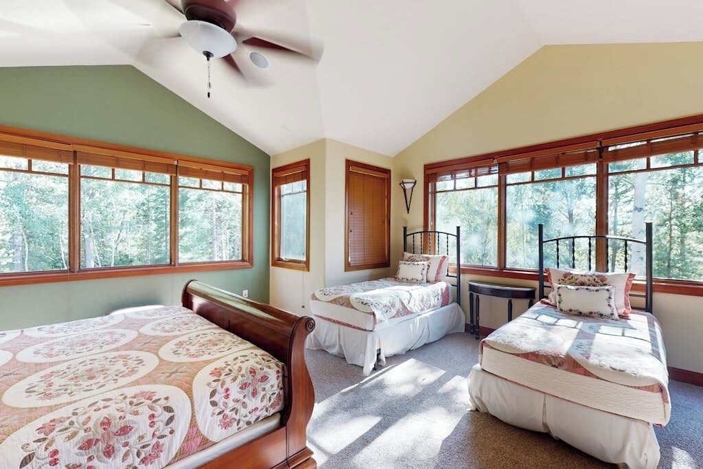 Una habitación con dos camas, ventilador de techo y ventanas amplias con vista a árboles.