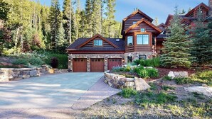 A large log cabin with multiple garage doors, surrounded by a stone retaining wall and a landscaped garden. - Luxury mountain estate w/ private hot tub, home theater, two W/Ds, & spa (Keystone)