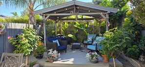 Eine überdachte Terrasse mit zwei blauen Stühlen und einem Tisch, umgeben von Topfpflanzen und einem Holzzaun.