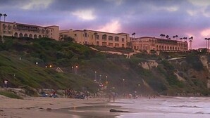 Un hotel frente a la playa con un edificio grande, una playa de arena y un acantilado rocoso.