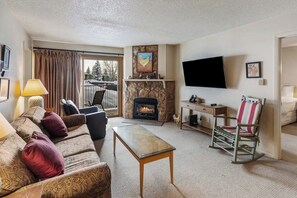 Ein Wohnzimmer mit Kamin, Fernseher, einer Couch, einem Schaukelstuhl und Blick ins Freie.