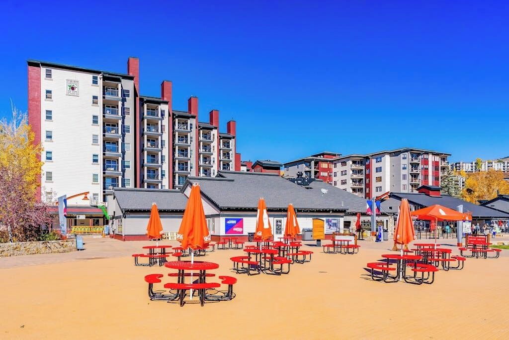 Un hotel con varios pisos, área de estar al aire libre con mesas y sombrillas, y un cielo azul despejado.