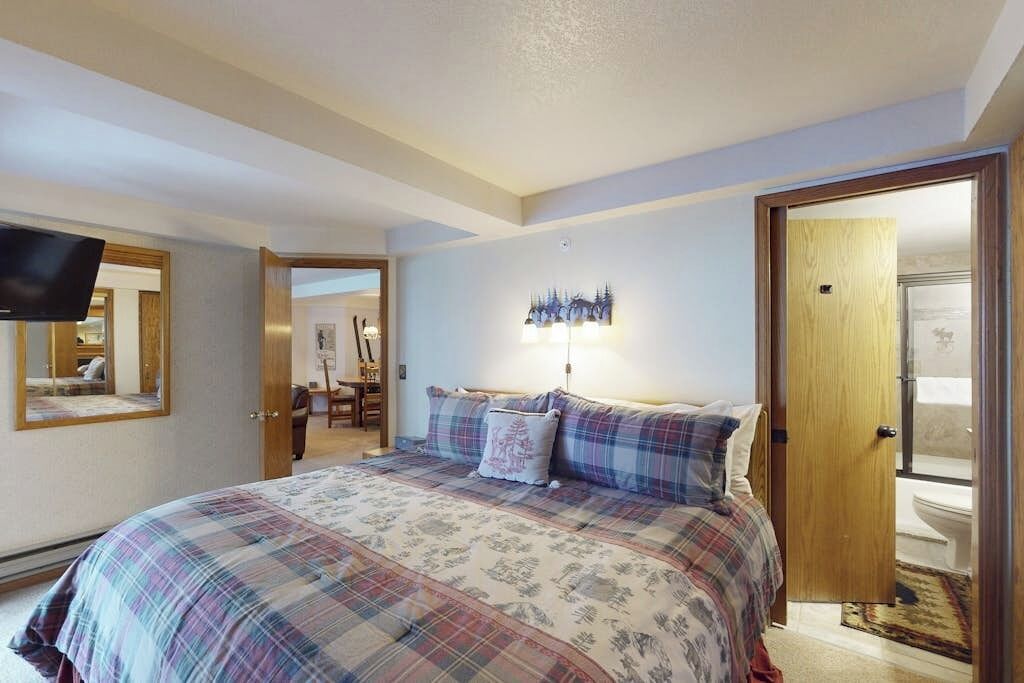 A bedroom with a bed, a television mounted on the wall, a door leading to a bathroom, and a mirror on the wall.
