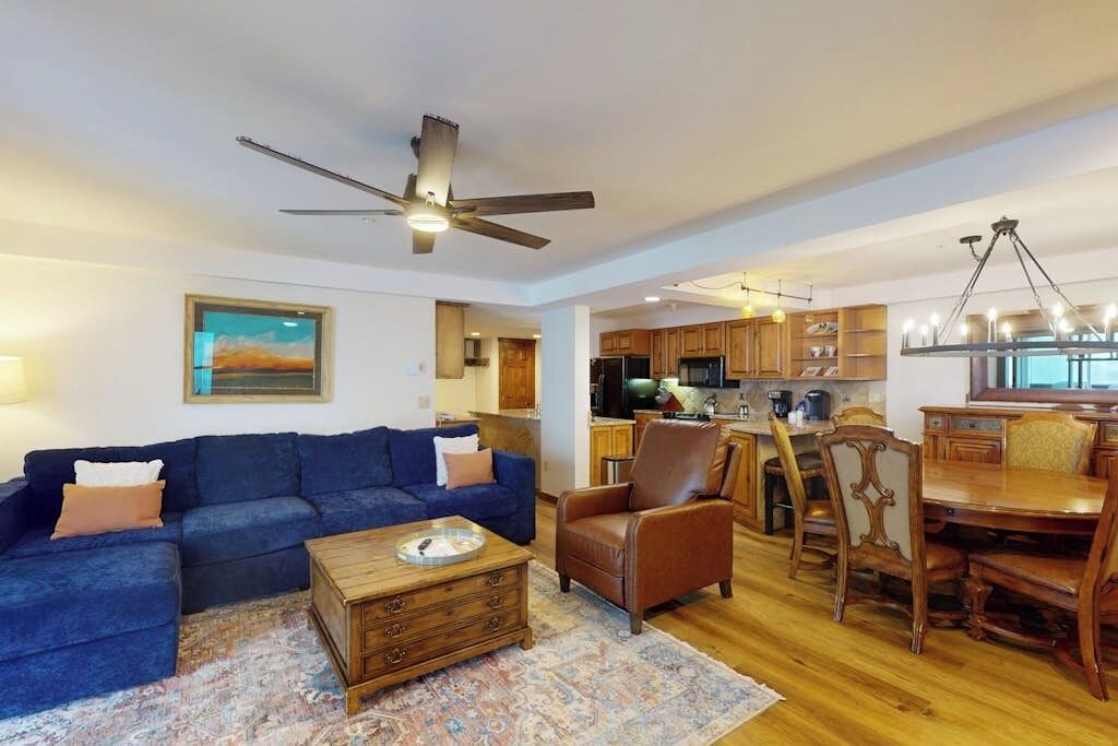 A living room with a blue sofa, a wooden coffee table, a brown leather chair, and a dining area with a wooden table and chairs.