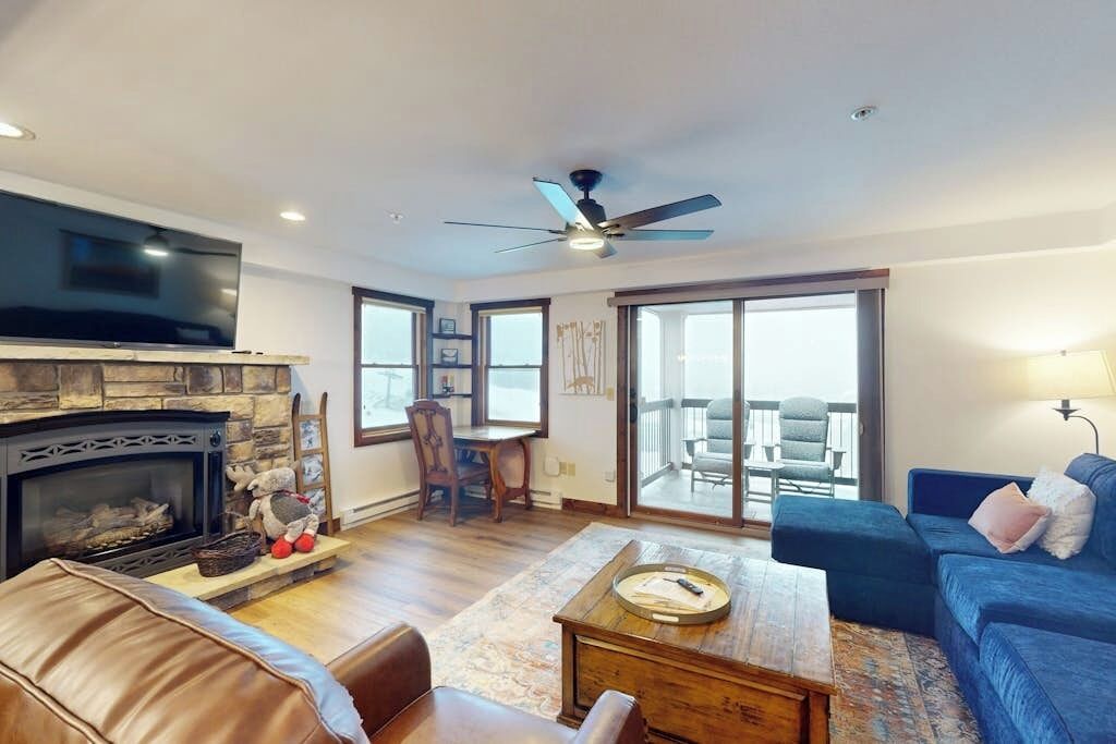 A living room with a fireplace, a television, a ceiling fan, a dining area with chairs, and a balcony with outdoor seating.