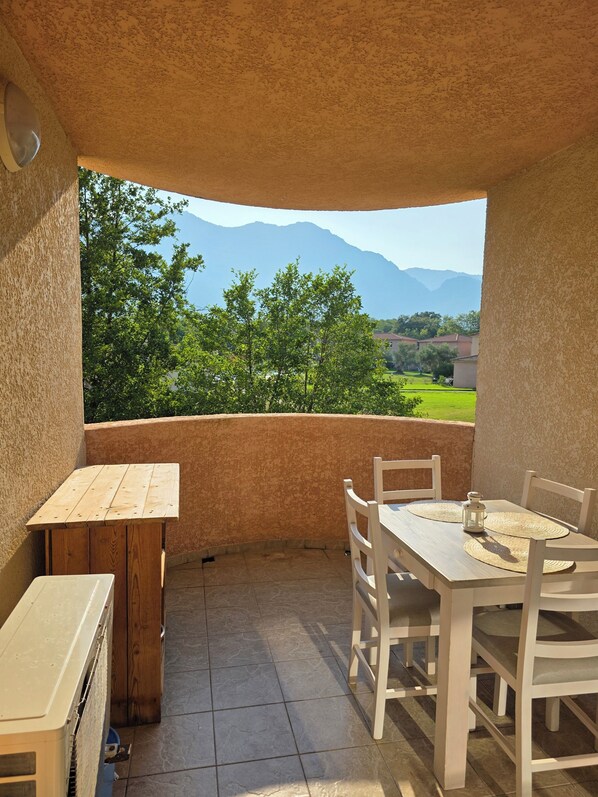 A covered outdoor patio with a table and chairs, a wooden table, and a view of green trees and mountains. - Corsica: sea, mountains and swimming pool (Santa-Maria-Poggio)