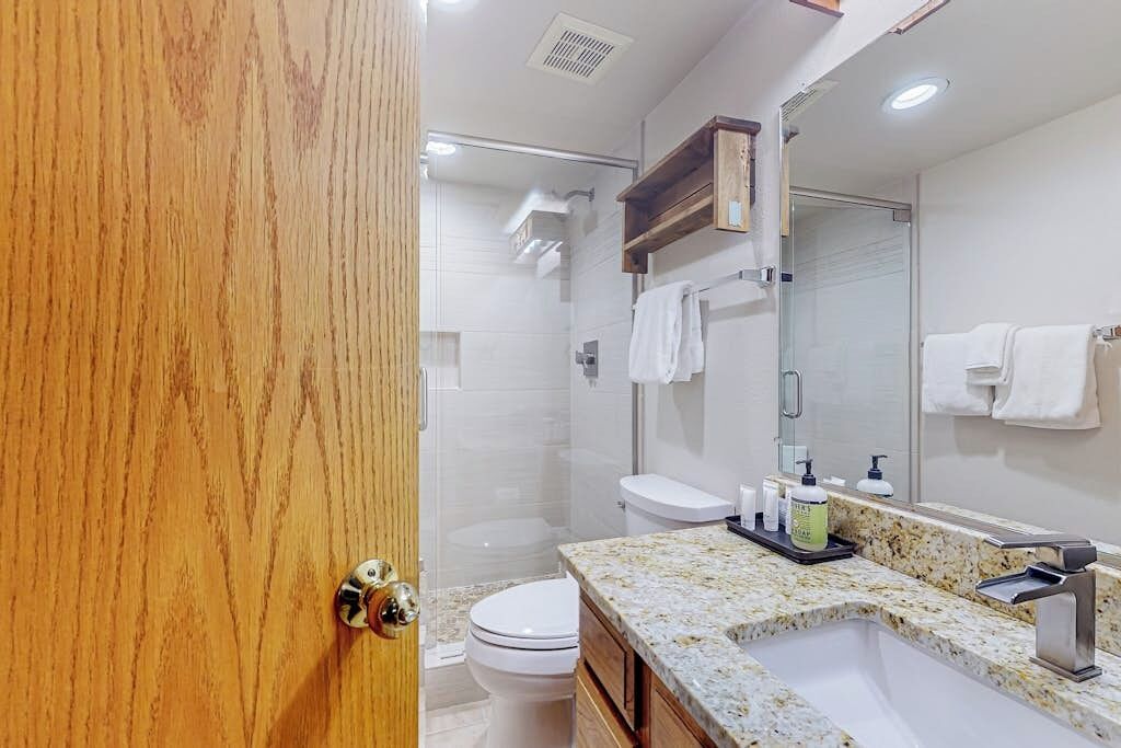 A modern bathroom with a wooden door, a granite countertop, a sink, a toilet, and a shower area.