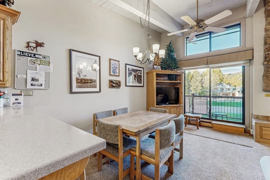 A dining area with a wooden table and chairs, a chandelier, and a view of the outdoors through large windows.