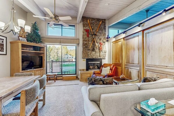A spacious living room with a fireplace, a ceiling fan, a large window, and a stone wall. - Ski-In/Out Top-Floor Condo w/Free WiFi, Fireplace, and Shared W/D, Hot Tub, Pool (Steamboat Springs)