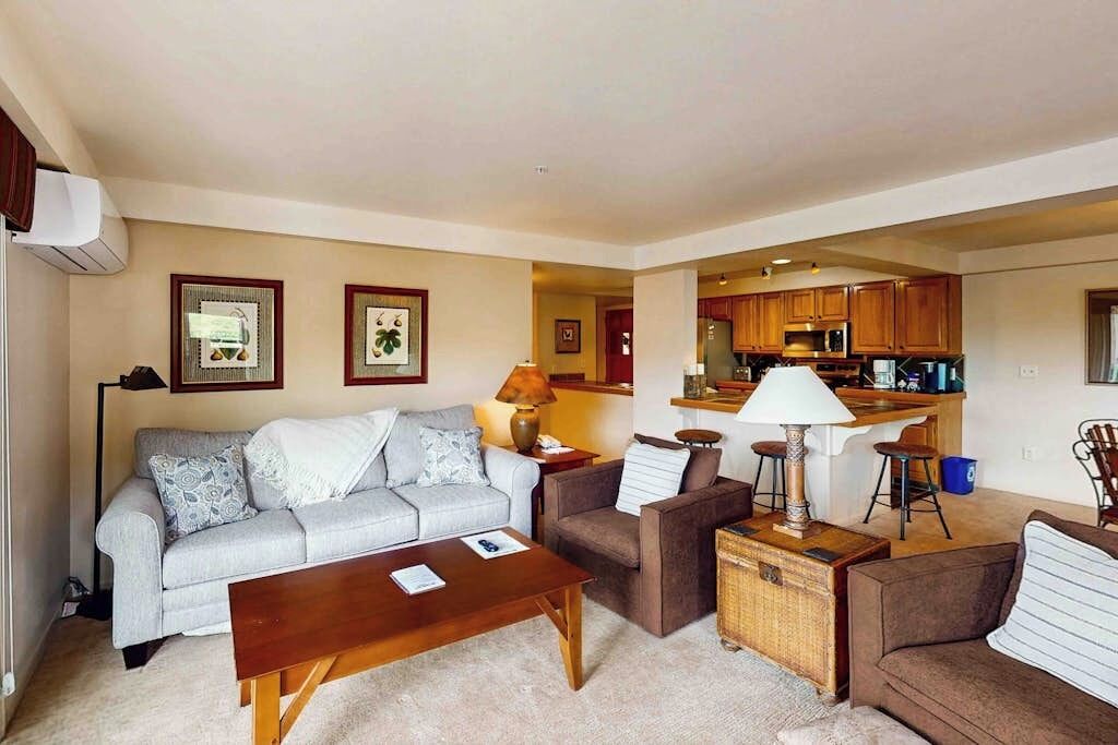 A living room with a sofa, armchair, coffee table, and a small kitchen area with a bar stool.
