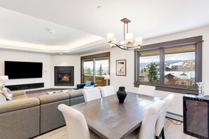 A modern living room with a fireplace, a large TV, and a dining area with a view of the outside. - Modern centrally located condo with pool, hot tub, gym & easy walk to lifts (Steamboat Springs)