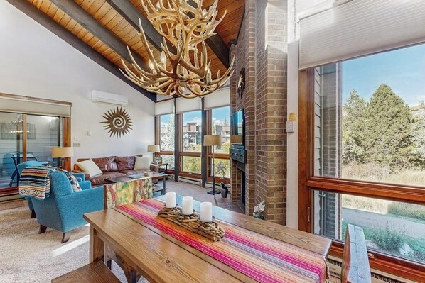 A spacious living room with a large wooden table, comfortable seating, a fireplace, and large windows offering a view of the outdoors. - Beautiful mountain condo with balcony, deck, hot tub & pool & washer/dryer (Steamboat Springs)