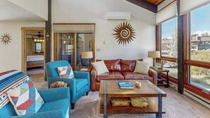 Un salón espacioso con un sofá de cuero, un sillón azul y una mesa de centro de madera. Una ventana grande ofrece vistas al exterior.