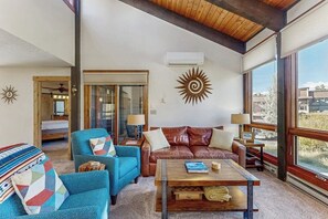 A spacious living room with a leather sofa, a blue armchair, and a wooden coffee table. There is a large window providing a view of the outside. - Beautiful mountain condo with balcony, deck, hot tub & pool & washer/dryer (Steamboat Springs)