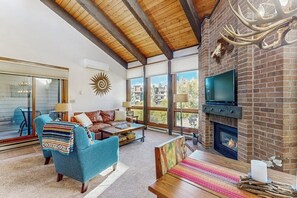 A spacious living room with a fireplace, a television, and a large window offering a view of the outside. - Beautiful mountain condo with balcony, deck, hot tub & pool & washer/dryer (Steamboat Springs)