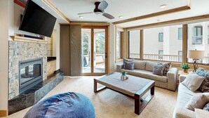 A living room with a flat-screen TV, a fireplace, a ceiling fan, a sofa, a coffee table, and large windows. - Meadows Normandy 1 | Spacious Mountain Condo, Fireplace, Shared Pool & Hot Tub, Winter Shuttle (Steamboat Springs)