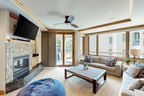 A living room with a flat-screen TV, a fireplace, a ceiling fan, a sofa, a coffee table, and large windows. - Conveniently located condo with fully kitchen, grill, shared hot tub & pool (Steamboat Springs)