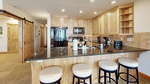 A modern kitchen with wooden cabinets, a granite countertop, stainless steel appliances, and three bar stools. - Meadows Normandy 1 | Spacious Mountain Condo, Fireplace, Shared Pool & Hot Tub, Winter Shuttle (Steamboat Springs)