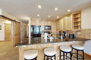 A modern kitchen with wooden cabinets, a granite countertop, stainless steel appliances, and three bar stools. - Conveniently located condo with fully kitchen, grill, shared hot tub & pool (Steamboat Springs)