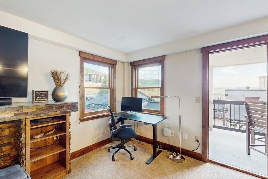Ein Zimmer mit Schreibtisch, Stuhl, Fernseher und Bergblick durch große Fenster.