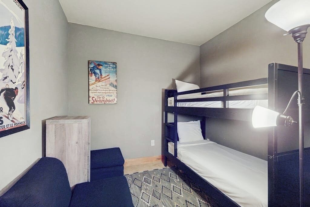 Habitación con literas, una lámpara, un cartel en la pared y un taburete de madera.