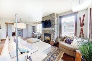 A living room with a stone fireplace, a television mounted above it, and a sofa with cushions. - Beautiful Mountain Condo Near Ski Resort w/Shared Pool/Hot Tub, WiFi, AC, & W/D (Steamboat Springs)