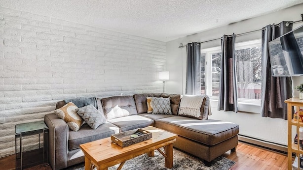 Ein modernes Wohnzimmer mit einer Eckcouch, einem Couchtisch aus Holz und einem großen Fenster mit Vorhängen.