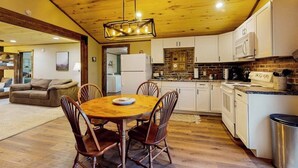 Un techo de madera, una mesa de comedor con sillas, una cocina con armarios blancos y un salpicadero de ladrillo, y una sala de estar con un sofá y una alfombra.