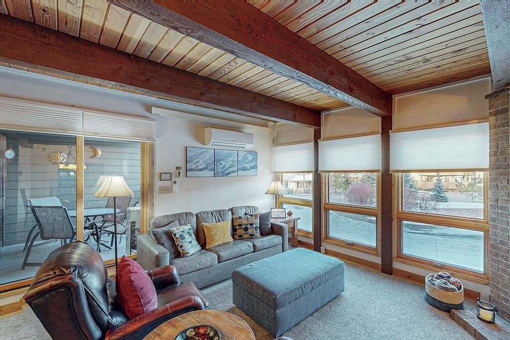 A cozy living room with a sofa, ottoman, and coffee table, large windows, and a fireplace.