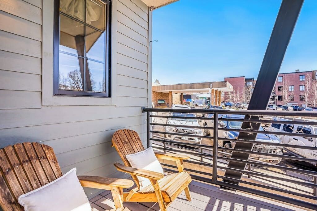 A balcony with two wooden Adirondack chairs and a window overlooking a parking lot and buildings.