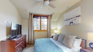 A bedroom with a bed, a TV on a dresser, a ceiling fan, a window with curtains, and a painting on the wall. - Dog-friendly Alpenglow townhome with gas fireplace, fast WiFi & great location; Alpen Glow 01 (Steamboat Springs)
