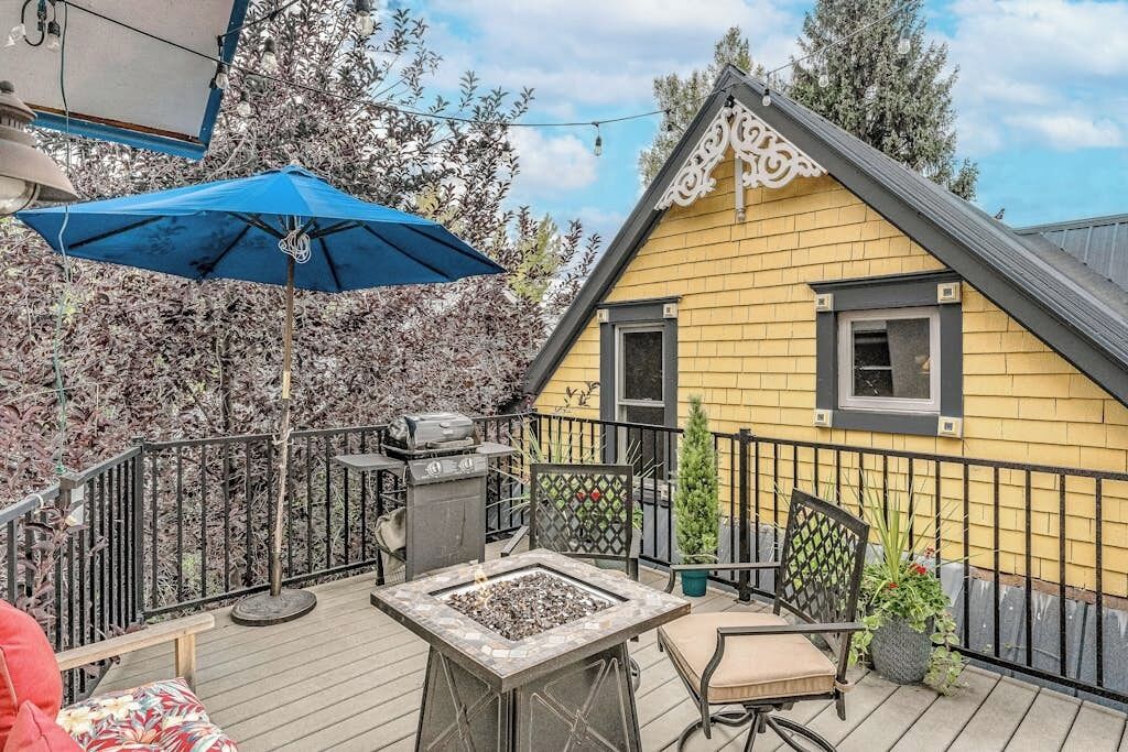 Una casa amarilla con techo negro, una terraza con un hogar de fuego, una parrilla y un paraguas azul.