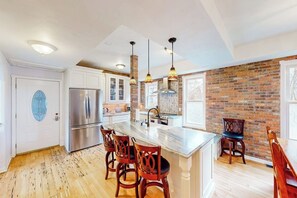 A kitchen with a central island, wooden chairs, a refrigerator, hanging pendant lights, and a brick wall. - Restored downtown home with front porch, mountain views, & washer/dryer (Steamboat Springs)