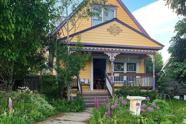 A yellow house with a porch, a mailbox, and a garden with purple flowers. - Restored downtown home with front porch, mountain views, & washer/dryer (Steamboat Springs)