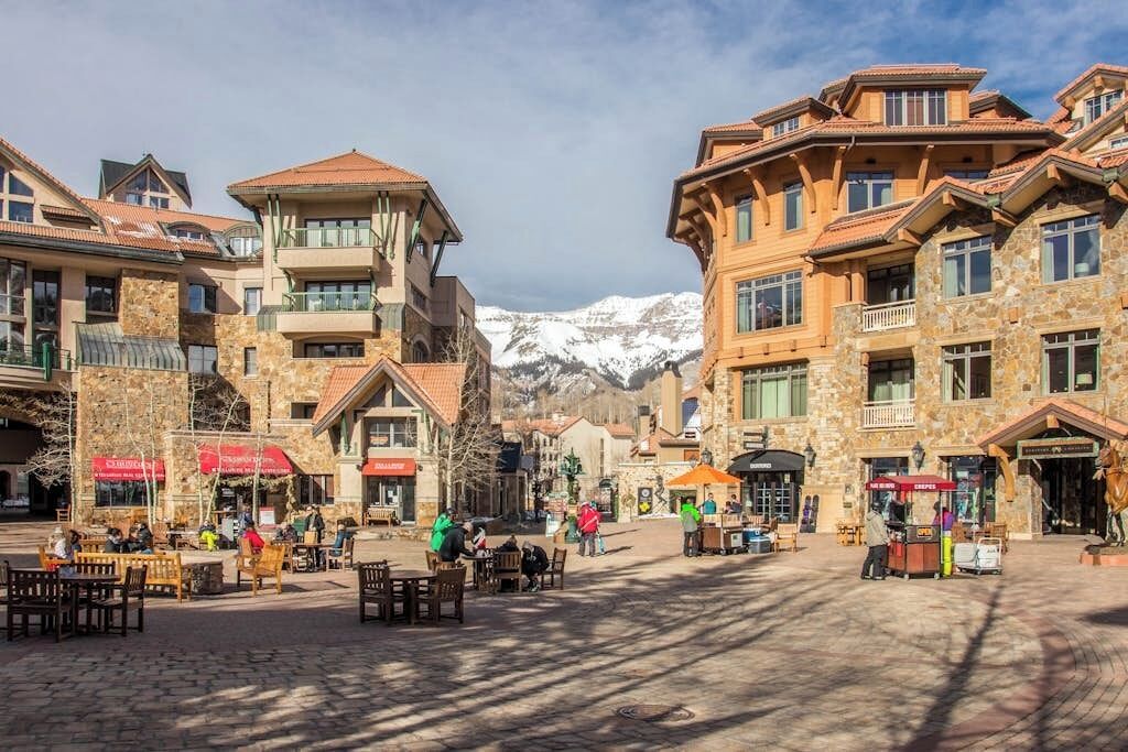 A town square with stone buildings, outdoor seating, and a mountain backdrop.